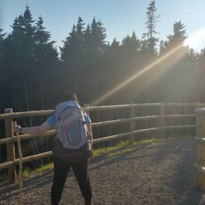 Photo of Michelle Brake holding awalking stick and wearing a backpack, looking towards a forest.