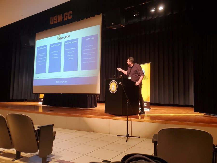 Eric Goforth stands at a podium on a stage. He is leaning forward and using a laser pointer to direct the audience's attention to a specific section of on a presentation that is being projected on a large banner next to him. The presentation currently shows four columns outlining the content of the presentation.