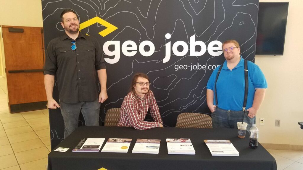 Eric Goforth, Brice Jones, and Danny Menikheim are standing in front of the GEO Jobe booth that the Mississippi Geospatial Conference. Flyers for various products and services are laid out in front of them. 