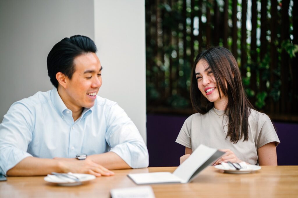 Image of two people seated at a table outside. The man on the left appears to be of East-Asian descent and is smiling while looking at the woman on his left. The woman is smiling, perhaps just about to laugh, while looking back at him. They appear to be mid-conversation.