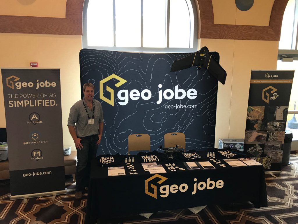Reed Davis stands next to the GEO Jobe booth at the APA conference. The booth is adorned with flyers and various swag items. There are also multiple drones on the table and around the booth.