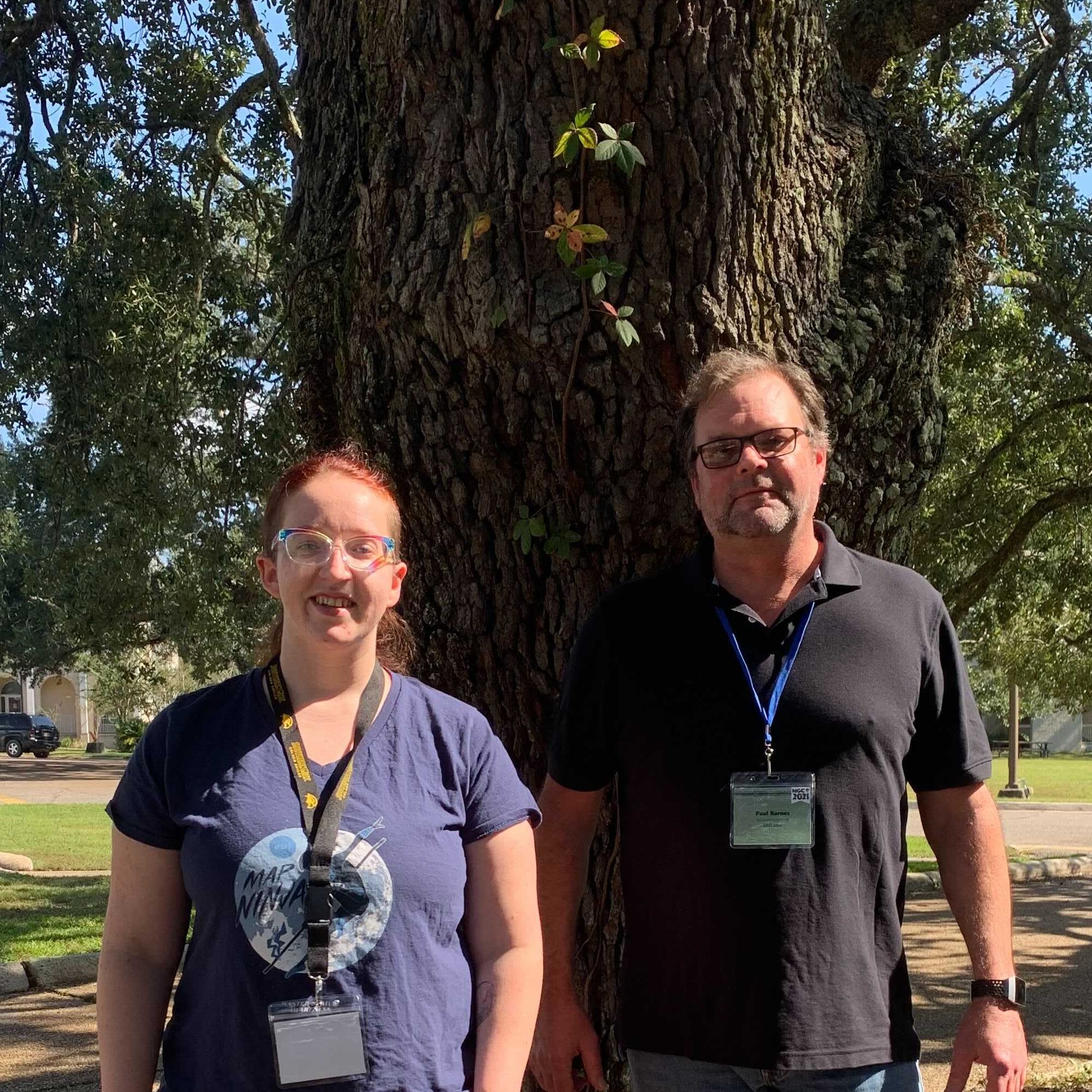 Courtney Menikheim and Paul Barnes stand before an oak tree at the Mississippi Geospatial Conference. Courtney is wearing glasses and a shirt that says "Map Ninja". Paul Barnes is wearing glasses and a black polo. They have just been re-elected as MAST's Secretary and Treasurer, respectively.