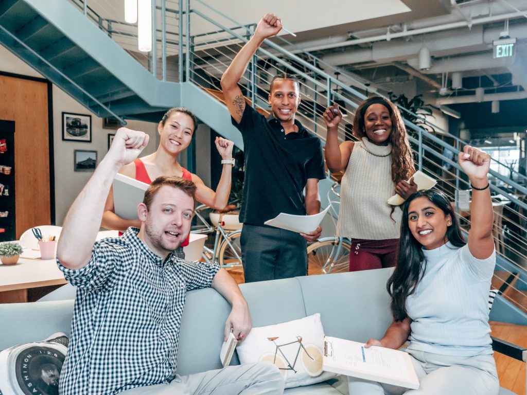 The same five people from the first image are now sitting and standing around a couch. They are all looking at the camera and smiling. All of them have an arm raised with a fist, indicating victory and celebrating an accomplishment.