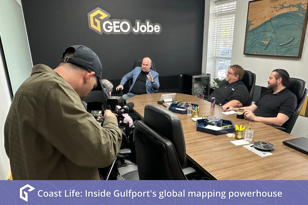 A film crew records a business interview at GEO Jobe in Gulfport, featuring three men discussing mapping technology in a modern conference room. Across the bottom of the photo is a solid purple bar overlaid with light contour lines and white stylized "G" logo. Next to it is text in white: "Coast Life: Inside Gulfport's global mapping powerhouse."